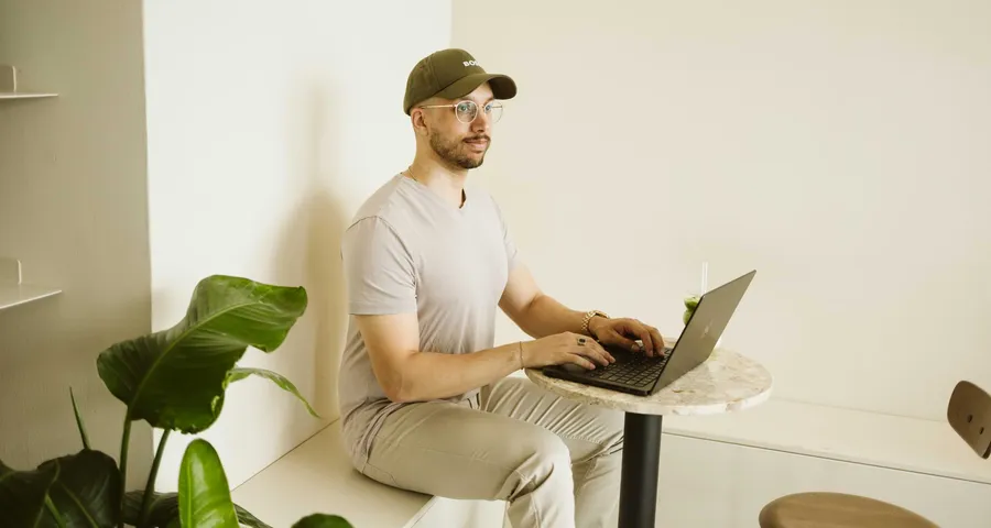 Dilshan Kelsen sitzt in einem Cafe mit seinem Laptop und einem Matcha-Latte und blickt in die Ferne.